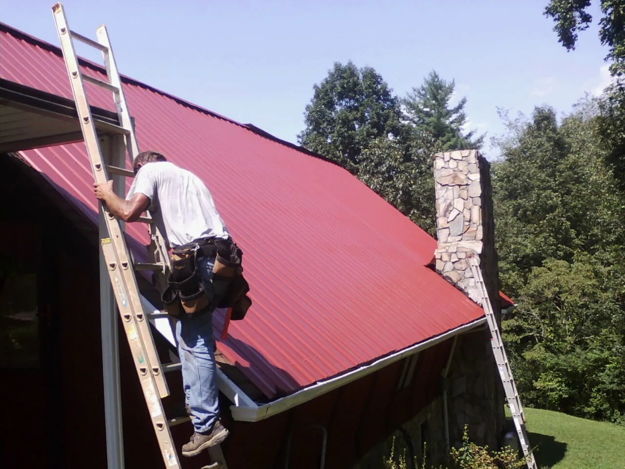 Roofer on a ladder beside a red metal roof and stone chimney — Soffit & Fascia Repair by Roofing Brothers in Filer